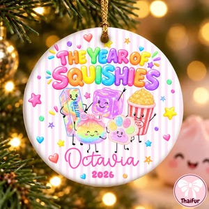 Puede incluir: Un adorno redondo de cerámica blanca con rayas verticales rosas que incluye el texto THE YEAR OF SQUISHIES, Octavia y 2026. El diseño presenta ilustraciones coloridas de una barra de mantequilla, un cubo, un cubo de palomitas y una huella.