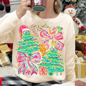 May include: Cream-coloured sweatshirt featuring a festive Christmas design. The graphic includes Christmas trees, bows, candy canes, and a gingerbread man. The design is in shades of pink, green, and gold.