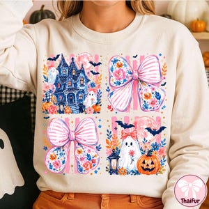 Puede incluir: Una sudadera de cuello redondo color crema con un estampado gráfico de cuatro paneles. El diseño incluye una casa encantada azul, dos grandes lazos rosas a rayas con detalles florales, un fantasma sosteniendo una flor, una calabaza, murciélagos y una luna llena.