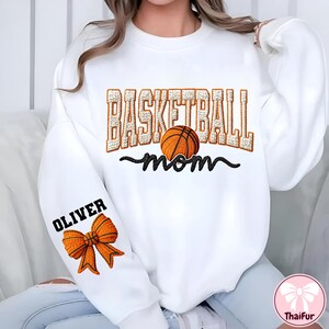 Puede incluir: Sudadera blanca con el texto "BASKETBALL mom" en naranja y negro, con un gr&aacute;fico de baloncesto. La manga tiene el nombre "OLIVER" encima de un gr&aacute;fico de lazo naranja. Un logotipo de ThaiFur est&aacute; en la esquina inferior derecha.