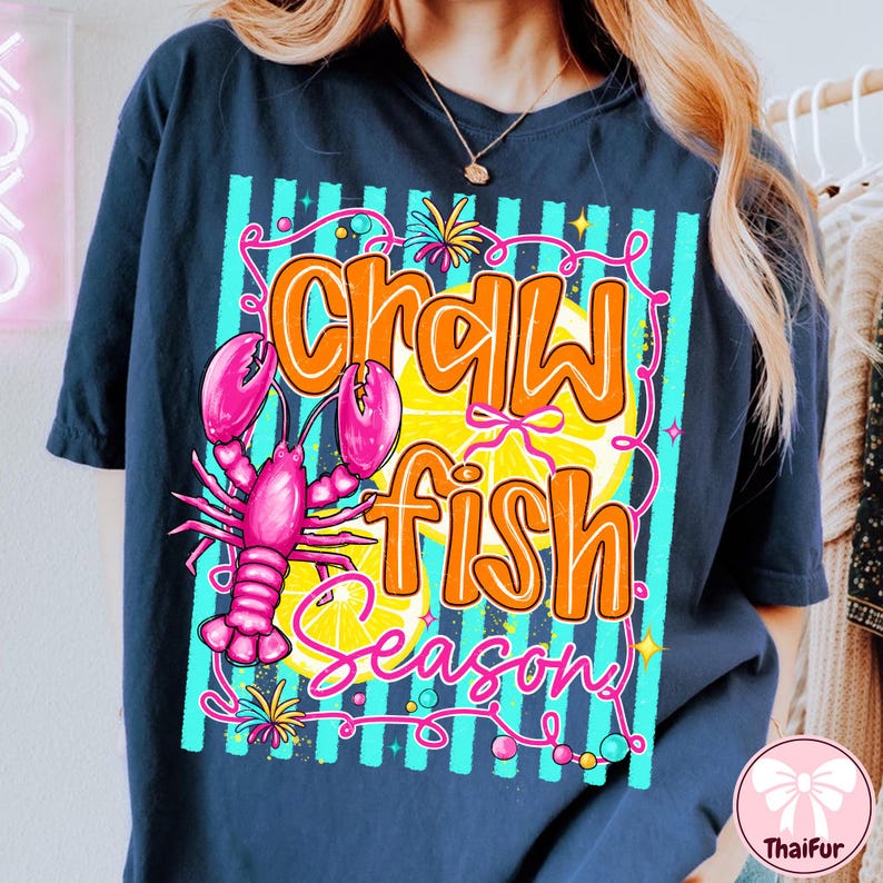 May include: Navy blue t-shirt with a colorful graphic. The design features the words "Crawfish Season" in orange and pink, a pink crawfish, and a yellow lemon slice. The background has teal and white vertical stripes.