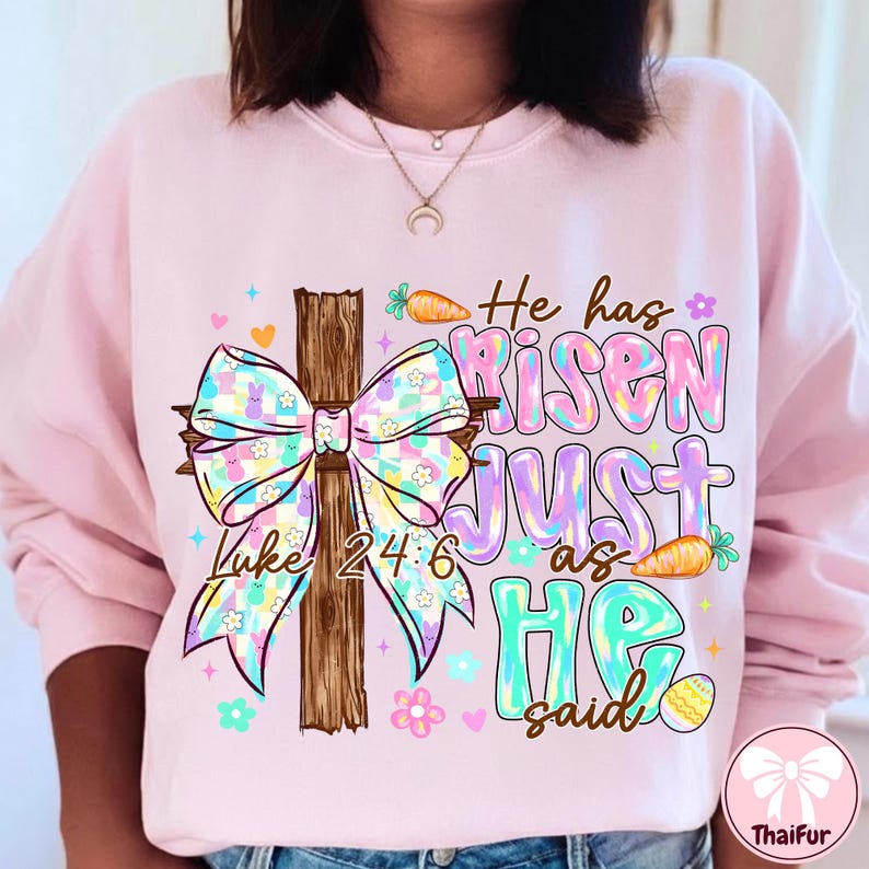 May include: A light pink sweatshirt with a colorful Easter-themed graphic. The design includes a wooden cross, a pastel bow, and the text "He has risen just as He said" with the Bible verse Luke 24:6.