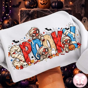 May include: A white sweatshirt featuring a colorful graphic with the text SPOOKY. The letters are decorated with mummy wrappings, red and blue patterns, and small illustrations of spider-man characters, ghosts, bats, pumpkins, and autumn leaves.