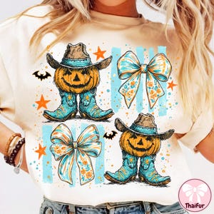 Op de afbeelding: Een crèmekleurig t-shirt met een grafische print van twee pompoenen met cowboyhoeden en turquoise cowboylaarzen, vergezeld door twee grote strikken met bloemmotieven, oranje sterren en kleine zwarte vleermuizen op een lichtblauwe achtergrond.
