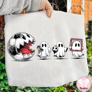 May include: White graphic tee featuring a soccer ball with a gaping mouth and three ghost figures. One ghost holds a soccer cleat, another a soccer ball, and the last is framed in a red rectangle. The design is in black and white.