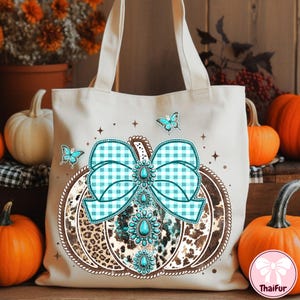 May include: Beige tote bag featuring a pumpkin design with a turquoise gingham bow and turquoise gemstone accents. The pumpkin has a leopard print pattern and is surrounded by butterflies. Perfect for fall.