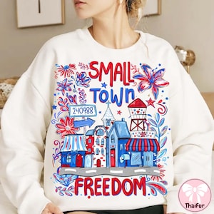May include: White sweatshirt with a colorful graphic design. The design features the words "SMALL TOWN FREEDOM" with illustrations of buildings, flowers, and a water tower in red, white, and blue.