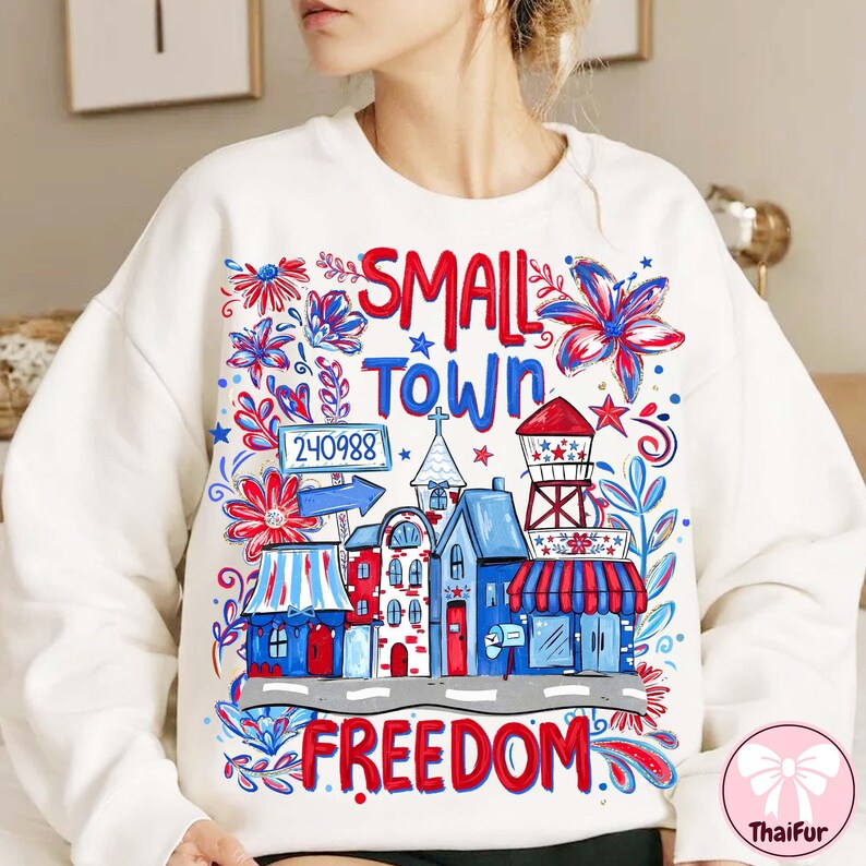 May include: White sweatshirt with a colorful graphic design. The design features the words "SMALL TOWN FREEDOM" with illustrations of buildings, flowers, and a water tower in red, white, and blue.