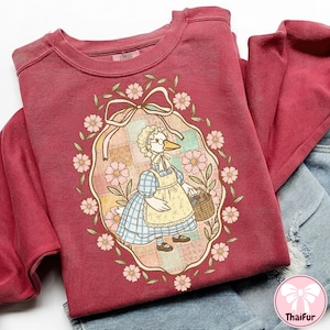 以下が含まれることがあります： ボンネットとエプロンを着てバスケットを持っているガチョウの気まぐれなイラストが特徴のマルーンのスウェットシャツ。 デザインは花のリースとリボンで囲まれています。 「COMFORT」という言葉がスウェットシャツの上部に印刷されています。