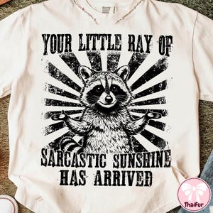 May include: Cream-colored t-shirt featuring a black and white graphic of a raccoon with outstretched arms, set against a sunburst design. The text reads "YOUR LITTLE RAY OF SARCASTIC SUNSHINE HAS ARRIVED."