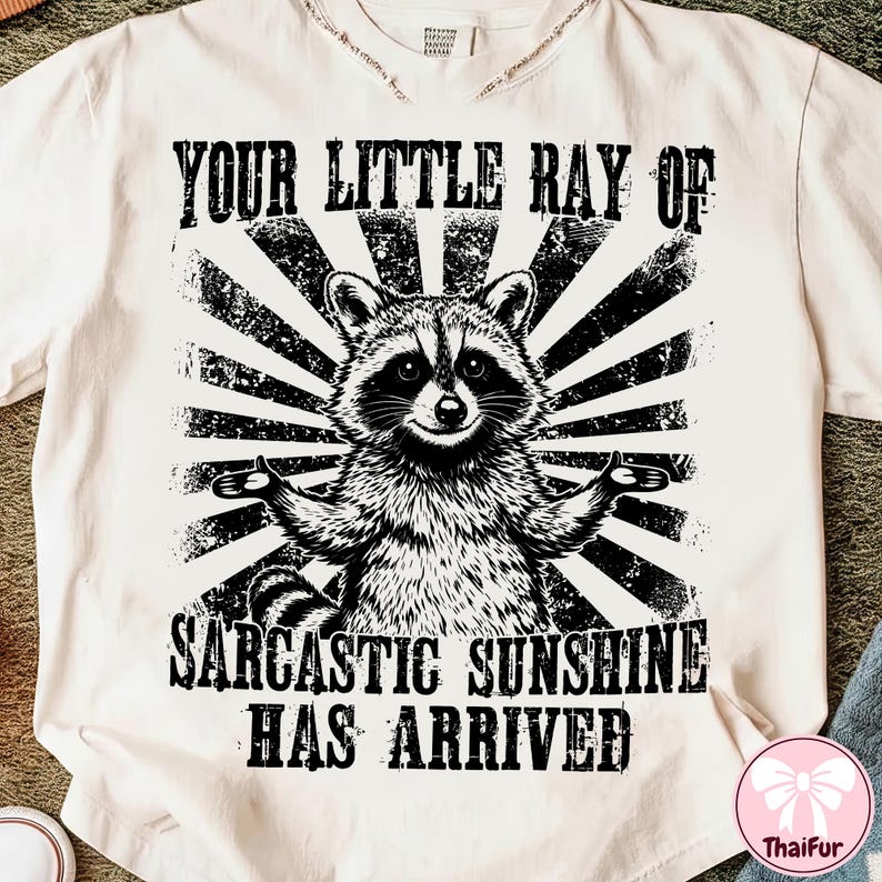 May include: Cream-colored t-shirt featuring a black and white graphic of a raccoon with outstretched arms, set against a sunburst design. The text reads "YOUR LITTLE RAY OF SARCASTIC SUNSHINE HAS ARRIVED."