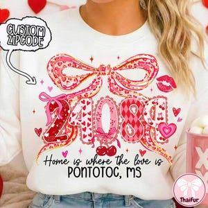 Op de afbeelding: Witte sweatshirt met een grote roze strik en de cijfers "24089" in een hartvormig ontwerp. De tekst "Home is where the love is PONTOTOC, MS" staat eronder gedrukt. Een "CUSTOM ZIPCODE" afbeelding staat linksboven.