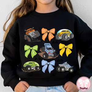 K&ouml;nnte beinhalten: Schwarzes Sweatshirt mit bunten Monstertruck-Grafiken und Schleifen. Die Trucks sind in Orange, Gelb, Gr&uuml;n und Grau gehalten und haben gro&szlig;e R&auml;der. Die Schleifen sind orange, gr&uuml;n, gelb und blau.