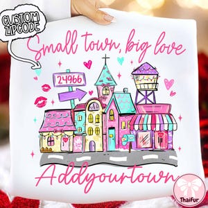 May include: White pillow with a colorful illustration of a small town. The design includes buildings, a water tower, and the text "Small town, big love" and "Add your town". A speech bubble says "Custom Zipcode".