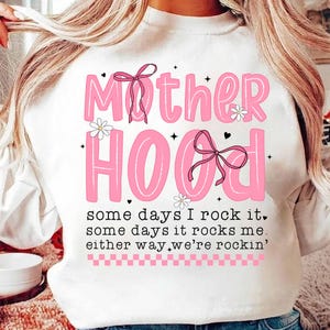 May include: White sweatshirt with pink lettering that reads "Mother Hood" with pink bows and flowers. The text below reads "some days I rock it, some days it rocks me, either way we're rockin'"
