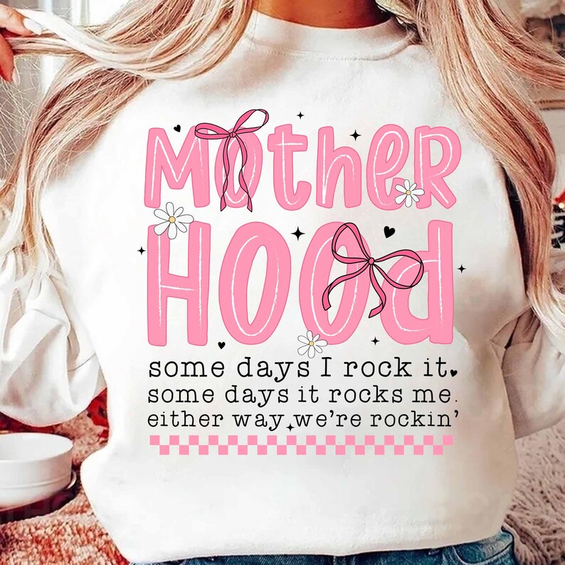 May include: White sweatshirt with pink lettering that reads "Mother Hood" with pink bows and flowers. The text below reads "some days I rock it, some days it rocks me, either way we're rockin'"