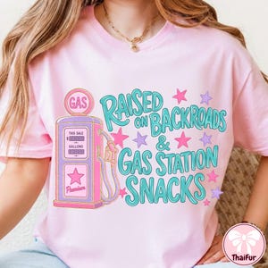 K&ouml;nnte beinhalten: Ein hellrosa T-Shirt mit einer Grafik einer alten Zapfs&auml;ule in Pink und Lila. Der Text RAISED ON BACKROADS & GAS STATION SNACKS ist in T&uuml;rkis gehalten und von kleinen pinken und lila Sternen umgeben. Das Design ist mittig auf der Vorderseite.