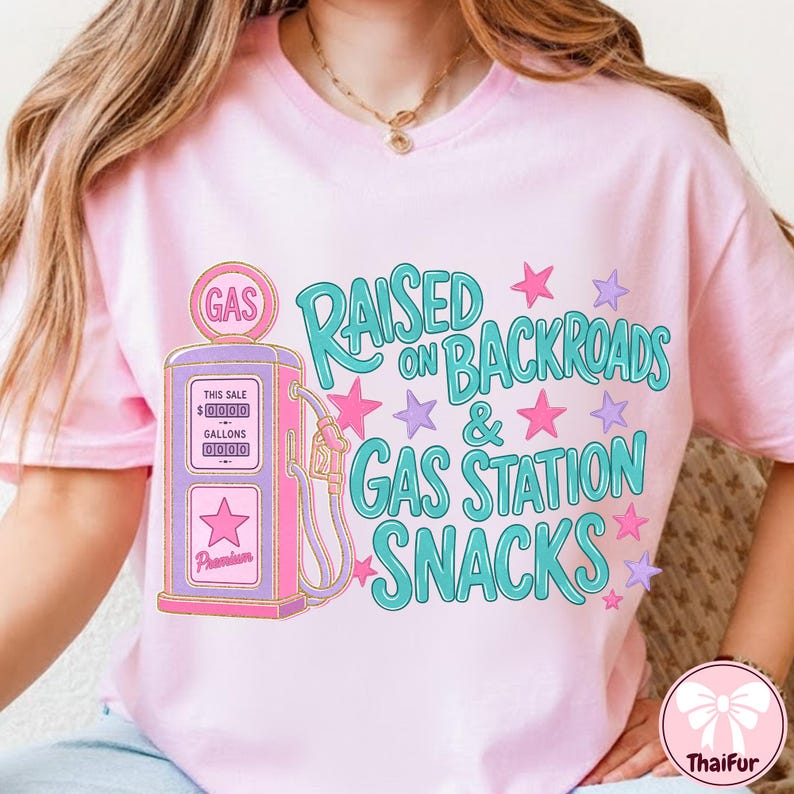 K&ouml;nnte beinhalten: Ein hellrosa T-Shirt mit einer Grafik einer alten Zapfs&auml;ule in Pink und Lila. Der Text RAISED ON BACKROADS & GAS STATION SNACKS ist in T&uuml;rkis gehalten und von kleinen pinken und lila Sternen umgeben. Das Design ist mittig auf der Vorderseite.