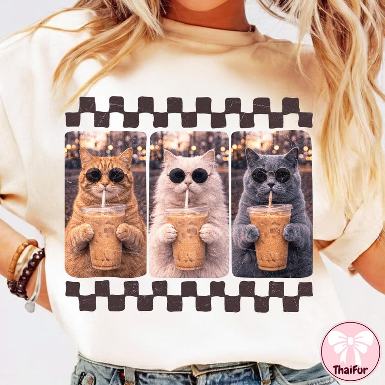 May include: Cream-colored t-shirt featuring a graphic of three cats wearing sunglasses and holding iced coffee. The cats are in a row, each with a different fur color: orange, white, and gray. A black and white checkered pattern frames the image.