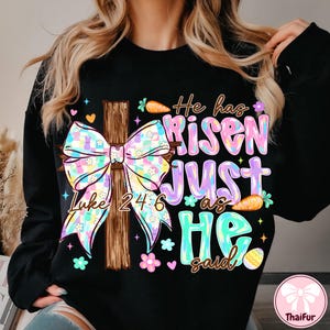 May include: Black sweatshirt with a colorful Easter design. Features a wooden cross with a pastel bow, the text "He has Risen JUST as HE said" and the Bible verse Luke 24:6. Includes carrots, flowers, and an Easter egg.