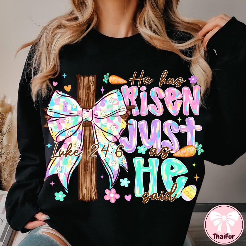 May include: Black sweatshirt with a colorful Easter design. Features a wooden cross with a pastel bow, the text "He has Risen JUST as HE said" and the Bible verse Luke 24:6. Includes carrots, flowers, and an Easter egg.