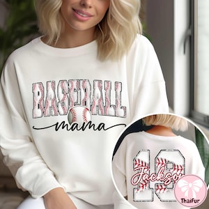 May include: Cream sweatshirt featuring the text "BASEBALL mama" in a baseball-themed design. The word "BASEBALL" is in a block letter style with red baseball stitching. A baseball graphic is centered below the word. The back has the number 19 and the name "Jackson".