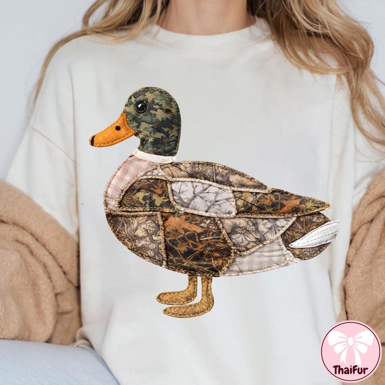 May include: Cream-colored sweatshirt featuring a large, appliqued duck design. The duck has a camouflage patterned head and body, with an orange beak and feet. The design is stitched onto the shirt.