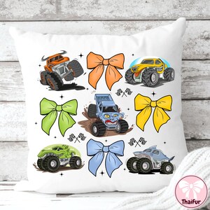 K&ouml;nnte beinhalten: Wei&szlig;es Kissen mit bunten Monster-Truck-Illustrationen und gro&szlig;en Schleifen. Die Trucks sind orange, gelb, gr&uuml;n und blau, mit schwarzen Reifen. Die Schleifen sind orange, gr&uuml;n, gelb und blau. Dekoratives Design.