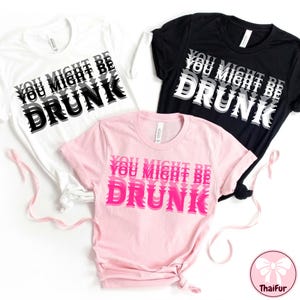 Puede incluir: Tres camisetas en blanco, negro y rosa, cada una con el texto "YOU MIGHT BE DRUNK" en una fuente estilizada en capas. La camiseta blanca tiene texto negro y gris, la camiseta negra tiene texto blanco y gris, y la camiseta rosa tiene texto rosa.