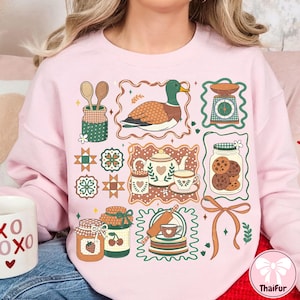 May include: Pink sweatshirt with a whimsical design featuring a duck, kitchen scale, cookies, jars of jam, and a teapot. The design includes wooden spoons, a rolling pin, and decorative frames. The overall theme is baking and kitchen decor.