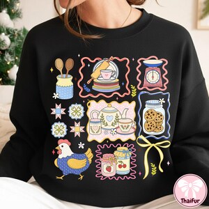 May include: Black sweatshirt with a vibrant, playful design showcasing baking-related illustrations. The design includes a teapot, cookies in a jar, a chicken, and other kitchen-themed graphics. The overall theme is centered around baking and cooking.