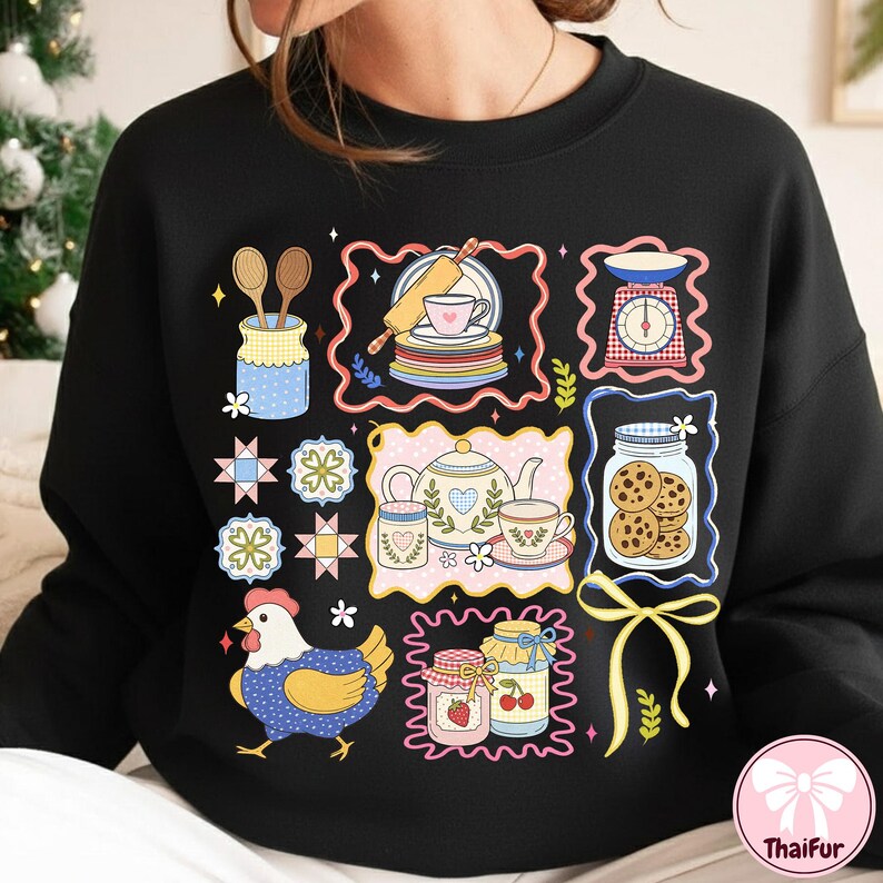 May include: Black sweatshirt with a vibrant, playful design showcasing baking-related illustrations. The design includes a teapot, cookies in a jar, a chicken, and other kitchen-themed graphics. The overall theme is centered around baking and cooking.