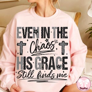 May include: A light pink sweatshirt with the text "EVEN IN THE Chaos HIS GRACE Still finds me" in black and white checkered and script lettering. The design includes crosses and stars.
