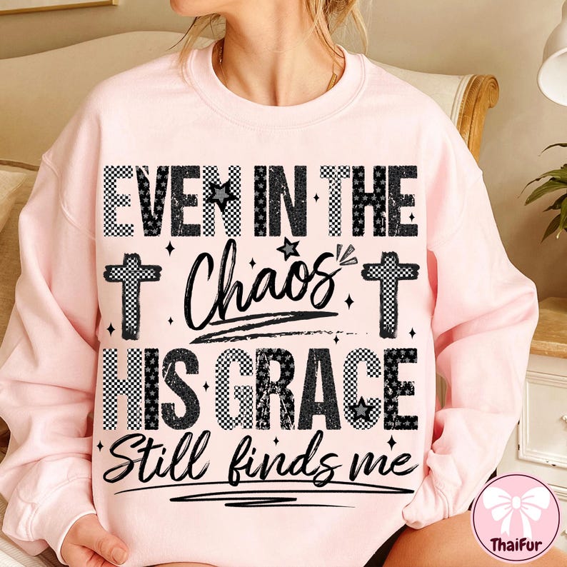 May include: A light pink sweatshirt with the text "EVEN IN THE Chaos HIS GRACE Still finds me" in black and white checkered and script lettering. The design includes crosses and stars.
