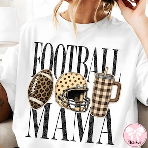 May include: A white t-shirt featuring the black text FOOTBALL MAMA. The design includes a leopard print football, a tan polka-dot football helmet, and a brown and white buffalo plaid tumbler with a straw, all arranged between the words.