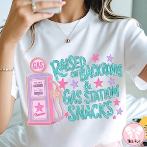 K&ouml;nnte beinhalten: Ein wei&szlig;es T-Shirt mit einer Grafik einer rosa und violetten Vintage-Zapfs&auml;ule. Der Text lautet RAISED ON BACKROADS & GAS STATION SNACKS in t&uuml;rkisfarbener Schrift. Das Design ist mit kleinen rosa und violetten Sternen verziert.