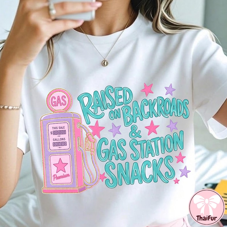 K&ouml;nnte beinhalten: Ein wei&szlig;es T-Shirt mit einer Grafik einer rosa und violetten Vintage-Zapfs&auml;ule. Der Text lautet RAISED ON BACKROADS & GAS STATION SNACKS in t&uuml;rkisfarbener Schrift. Das Design ist mit kleinen rosa und violetten Sternen verziert.