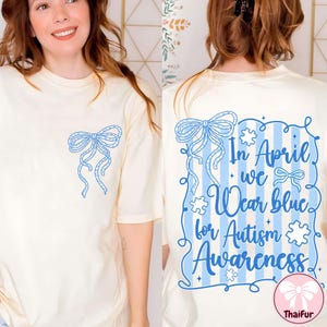 May include: Cream-colored t-shirt with a blue bow design on the front and back. The back of the shirt features the text "In April we Wear blue for Autism Awareness" in blue script and a blue bow design. The shirt is a short-sleeved, casual style.