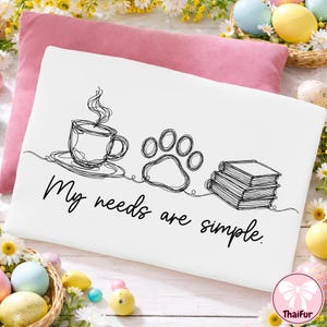 May include: White pillowcase with a black line drawing of a coffee cup, paw print, and books. The text "My needs are simple" is written below the drawings. A pink pillow is behind the pillowcase, with Easter decorations in the background.