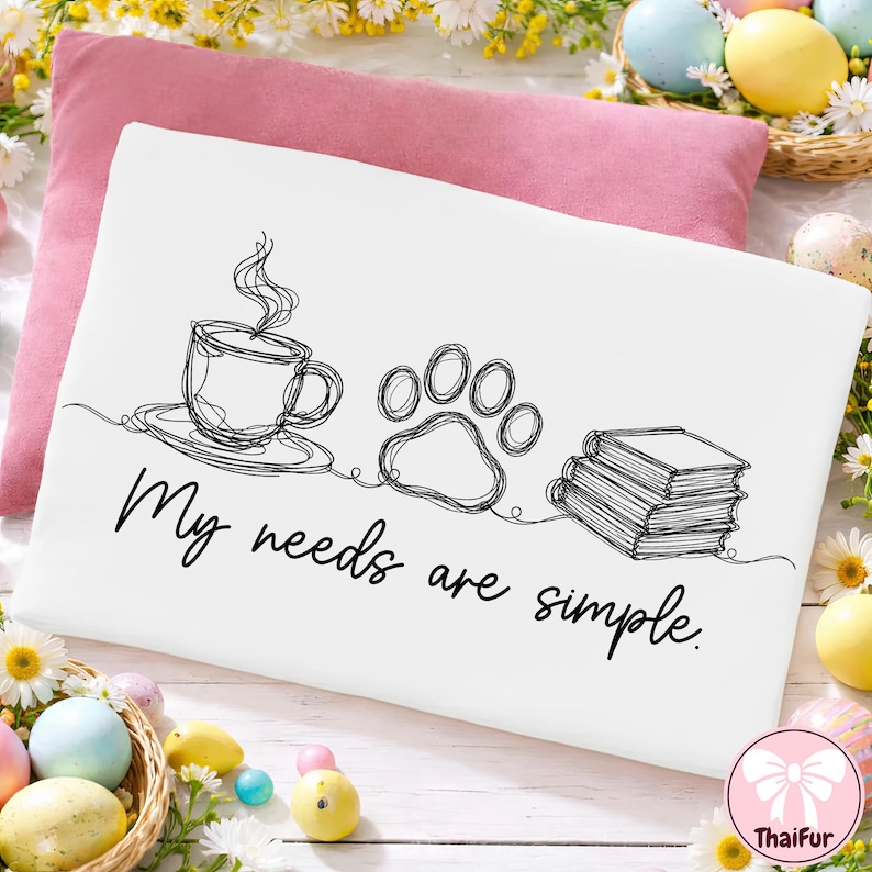 May include: White pillowcase with a black line drawing of a coffee cup, paw print, and books. The text "My needs are simple" is written below the drawings. A pink pillow is behind the pillowcase, with Easter decorations in the background.
