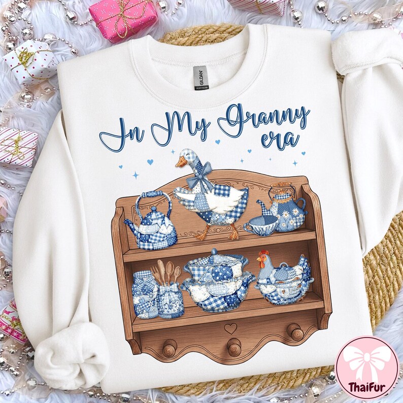 K&ouml;nnte beinhalten: Wei&szlig;es Sweatshirt mit dem Text "In My Granny era" in blauer Schrift. Die Grafik zeigt ein Holzregal mit blau-wei&szlig; karierten K&uuml;chenutensilien, einer Gans und einem Huhn. Das Design hat eine Vintage- und Landhaus&auml;sthetik.
