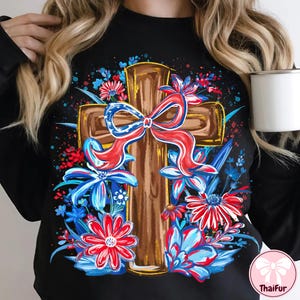 May include: Black sweatshirt featuring a brown cross design adorned with a red, white, and blue bow and floral accents. The flowers are in shades of red, white, and blue. The design is centered on the front of the shirt.