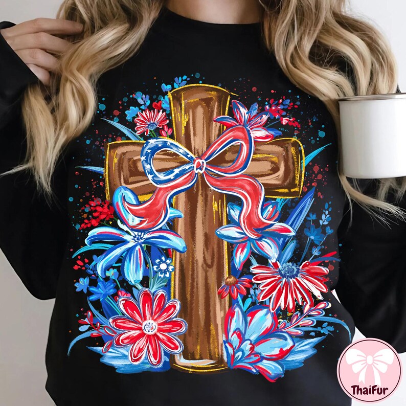 May include: Black sweatshirt featuring a brown cross design adorned with a red, white, and blue bow and floral accents. The flowers are in shades of red, white, and blue. The design is centered on the front of the shirt.