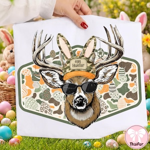 May include: White tote bag featuring a deer head wearing sunglasses and a camouflage hat that says "Egg Hunter." The deer has bunny ears and is surrounded by Easter eggs. The background has an orange, green, and brown camouflage pattern.