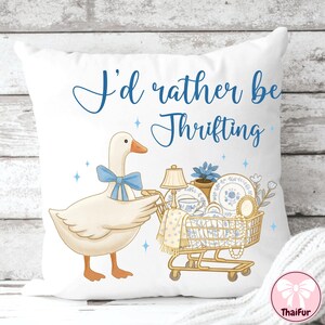 May include: White throw pillow with a whimsical design. A cartoon goose with a blue bow pushes a shopping cart filled with vintage dishes. The text "I'd rather be Thrifting" is written in blue script.
