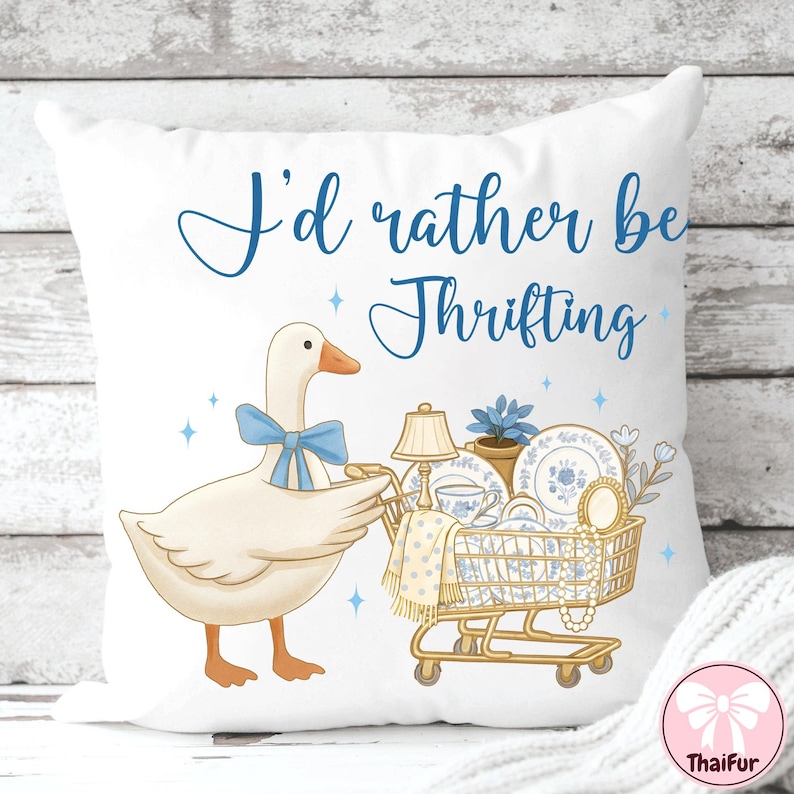 May include: White throw pillow with a whimsical design. A cartoon goose with a blue bow pushes a shopping cart filled with vintage dishes. The text "I'd rather be Thrifting" is written in blue script.