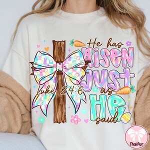 May include: Cream-colored t-shirt with a colorful Easter-themed graphic. The design features a wooden cross with a pastel bow, the text "He has Risen Just as He said" and the Bible verse Luke 24:6. Includes carrots, flowers, and an Easter egg.