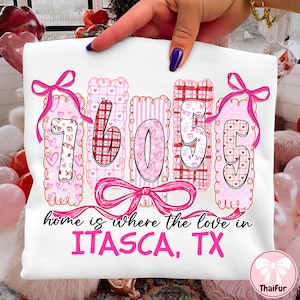 Puede incluir: Camiseta blanca con los números "76055" en rosa, decorada con corazones y lazos. El texto "home is where the love in ITASCA, TX" está impreso debajo. El diseño es sostenido por una persona.