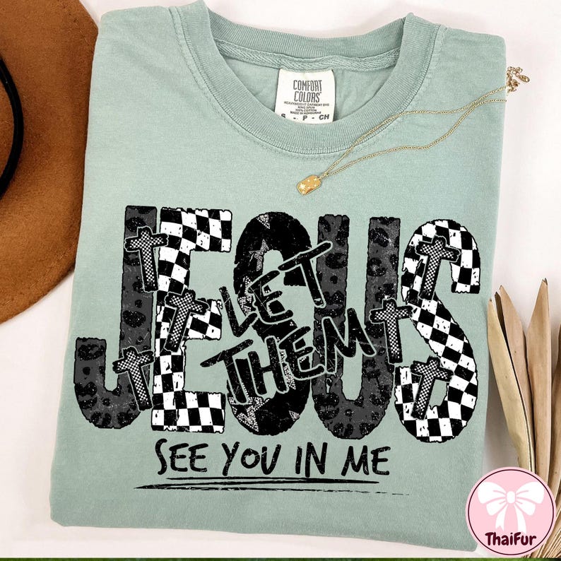 May include: A sage green t-shirt with the words "JESUS LET THEM SEE YOU IN ME" in a black and white checkered and leopard print design. A gold necklace is draped across the shirt.