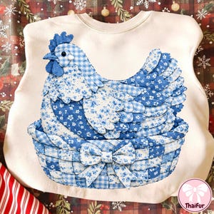 Op de afbeelding: Een crèmekleurige sweatshirt met een blauw en wit stoffen kippenontwerp. De kip zit in een mand met een strik. De kip en de mand zijn gemaakt van stof met patronen. Het ontwerp is gecentreerd op de voorkant van de trui.
